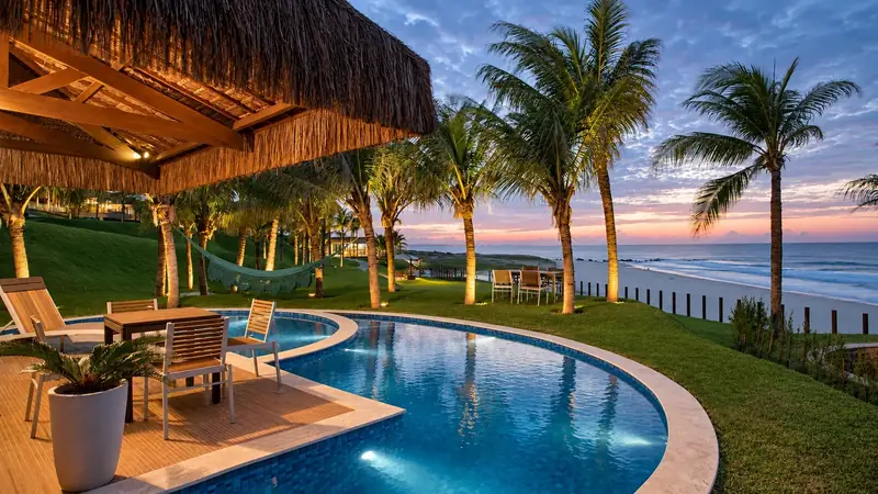 Piscina iluminada entre coqueiros, de frente para a praia, ao entardecer